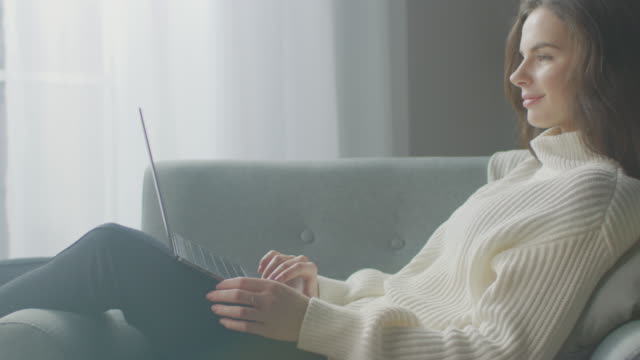 Schöne-junge-Frau-arbeitet-auf-Laptop-Computer,-während-auf-dem-Stuhl-sitzen.-Sinnliche-Mädchen-tragen-Pullover-funktioniert-auf-Notebook;-Studien,-Surfs-Internet,-Nutzt-Social-Media-beim-Entspannen-in-Cozy-Apartment