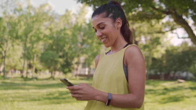 Dolly-shot-of-a-smiling-young-fit-woman-using-her-mobile-phone-looking-away-in-the-park
