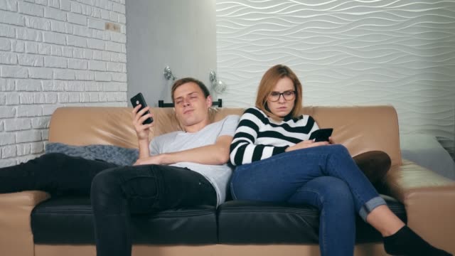 Attraktivejunges-Paar-sitzt-zurück-zu-Hause-auf-der-Couch-besessen-von-Smartphones.-Paar-mit-Handys-ignorieren-einander-als-Fremde,-Kommunikationsprobleme-und-soziale-Netzwerksucht