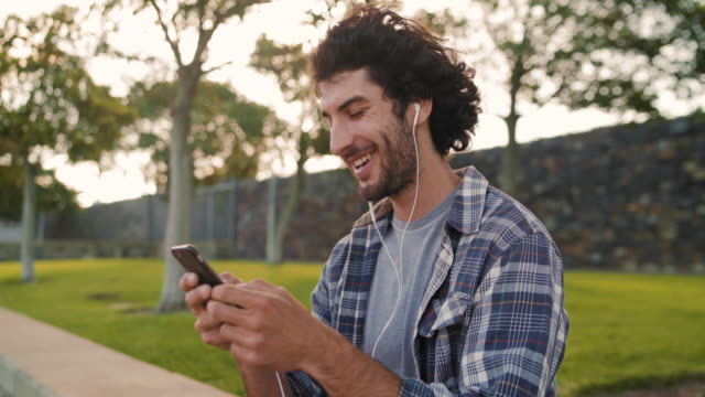 Retrato-de-un-joven-guapo-feliz-usando-auriculares-en-los-oídos-disfrutando-de-mensajes-de-texto-en-el-teléfono-inteligente-en-el-parque---hombre-feliz-usando-su-móvil-en-el-parque