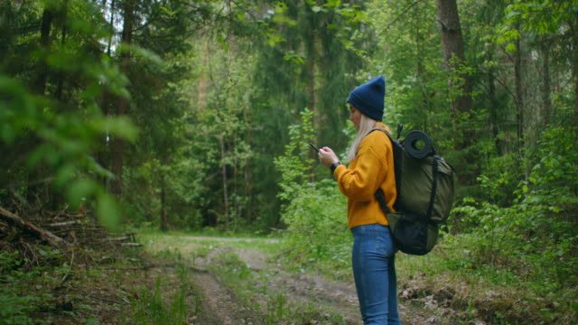 Cámara-lenta:-Joven-excursionista-usando-su-teléfono-inteligente-mientras-disfruta-de-su-fin-de-semana-en-el-bosque.-Mujer-viajera-con-mochila-caminando-por-el-camino-del-bosque-mirando-el-teléfono-y-mapa-en-madera-verde