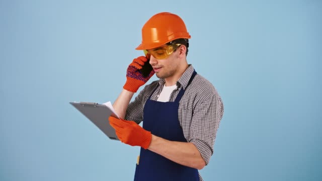 Young-male-in-safety-clothes,-hard-hat,-protective-goggles,-gloves.-Talking-by-cellphone,-looking-at-papers-on-clipboard.-Posing-on-blue-background