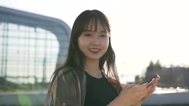 Gut-aussehende-glückliche-asiatische-Brünette-mit-langen-Haaren-halten-ihr-Handy-und-Blick-auf-die-Kamera-in-der-Nähe-des-modernen-Flughafens