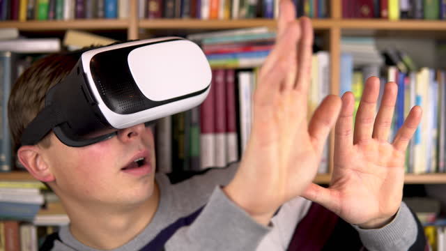 Junger-Mann-in-VR-Brille-in-der-Bibliothek.-Ein-Mann-mit-VR-Helm-auf-dem-Kopf-untersucht-und-berührt-die-virtuelle-Realität.-Im-Hintergrund-sind-Bücher-über-Bücherregale.-Buchbibliothek.