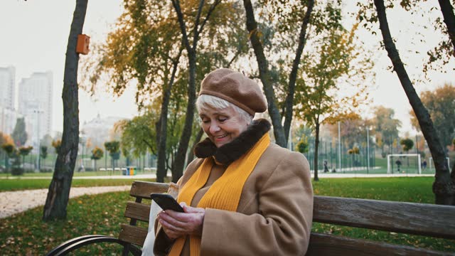 Gray-haired-woman-typing-message-on-mobile-phone-and-smiling-while-sitting-on-wooden-bench-in-autumn-city-park
