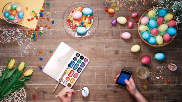 Man-painting-easter-egg-and-using-smartphone-on-table-decorated-with-easter-eggs.-Top-view