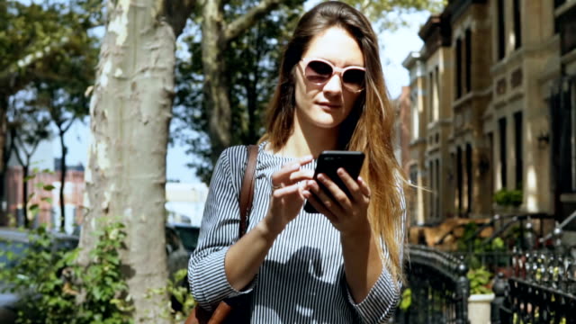 Portrait-of-young-beautiful-woman-in-sunglasses-walking-outside-in-sunny-summer-day-and-using-smartphone.-Slow-motion