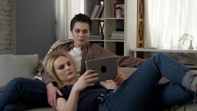 Lesbian-couple-is-resting-on-the-couch,-using-tablet-computer,-speaking,-showing-sign-no-by-shaking-head-60-fps