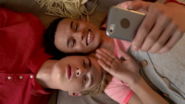 Happy-international-gay-couple-is-lying-on-the-couch-and-making-selfies-on-a-smartphone.-Homeliness,-LGBT-lovers,-happy-gay-family-concept,-laughing.-Say-hallo.-Video-chat.-Top-shoot-60-fps