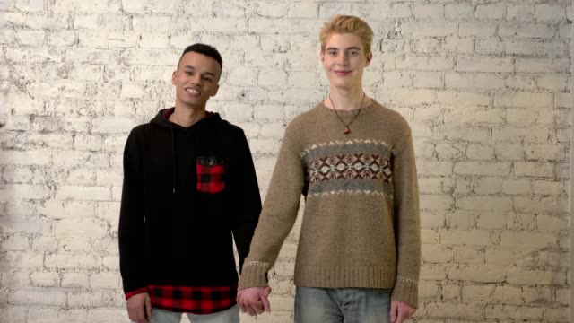 Internationalen-schwules-Paar-Hand-in-Hand,-Lächeln,-weiße-Mauer-im-Hintergrund.-LGBT-Liebe,-junges-Paar,-Liebhaber,-Homosexualität-Konzept.-60fps
