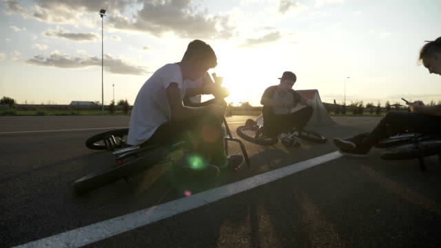 Relaxed-teenage-bikers-friends-taking-a-break-after-a-biking-training-on-circuit-using-smartphones-surfing-on-internet-and-resting-at-sunset