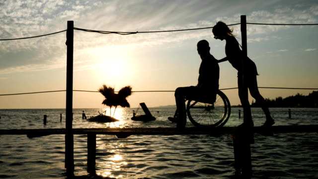 formschöne-Frau-mit-Männern-Behinderten-im-Rollstuhl-auf-Steg-am-Damm-gegen-den-Himmel-im-Abendrot