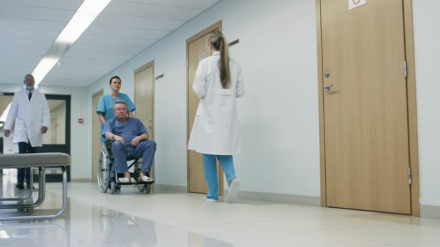 Arzt-spricht-mit-ihnen-zwar-im-Krankenhaus-Flur,-Krankenschwester-schiebt-älteren-Patienten-im-Rollstuhl,-Tablet-Computer-verwenden.-Sauberes,-neues-Krankenhaus-mit-medizinischen-Fachpersonal.
