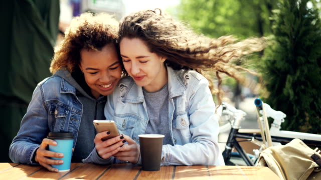 Attractive-young-ladies-Caucasian-and-African-American-are-using-smartphone-looking-at-screen-and-laughing-sitting-in-outdoor-cafe-at-table-together.