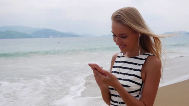 Young-beautiful-slim-woman-with-long-blonde-hair-in-black-and-white-dress-standing-on-the-coast-and-using-smartphone-over-background-at-storm-on-the-sea..-Girl-on-the-beach-touching-screen-and-smile.