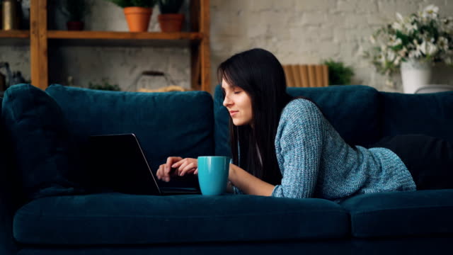 Attractive-young-woman-in-casual-clothes-is-typing-on-laptop-and-smiling-while-lying-on-sofa-in-loft-style-studio.-Modern-technology-and-millennials-concept