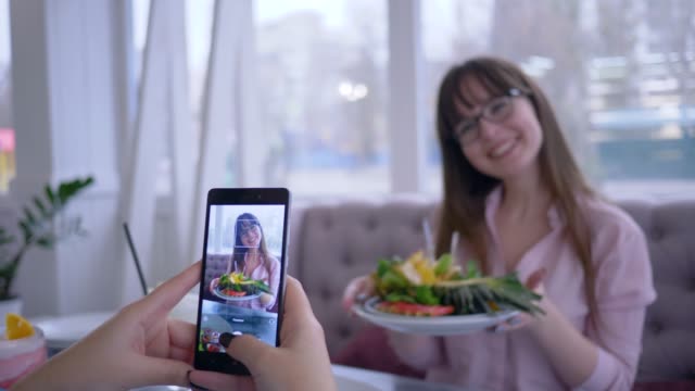 healthy-diet-and-blogging,-cheerful-girl-in-eyeglasses-posing-with-fresh-fruits-on-plate-for-girlfriend-who-takes-photo-on-mobile-phone-for-social-networks-in-cafe