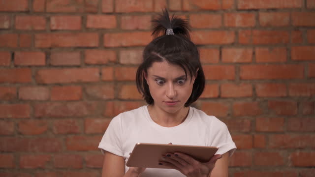 A-young-girl-in-shock-while-using-tablet-device-standing-isolated-against-a-brick-wall-background.