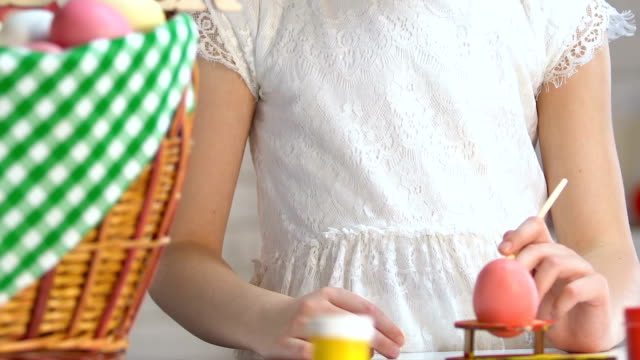 Little-girl-decorating-egg-with-paint,-Easter-greeting-basket-standing-on-table