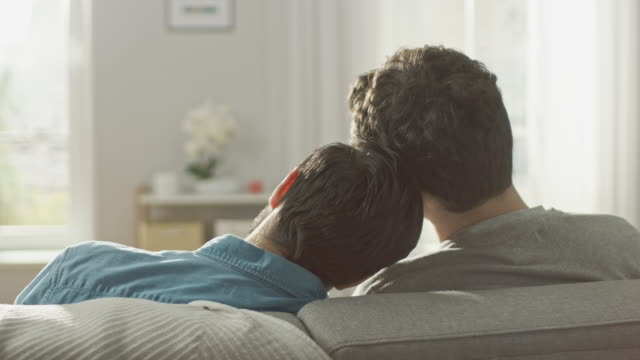 Back-Shot-of-Cute-Male-Queer-Couple-Sitting-on-a-Sofa-and-Putting-Their-Heads-Together.-They-are-Casually-Dressed-in-Jeans-and-Sweater.-Room-has-Modern-Interior-and-It's-Bright-and-Sunny.