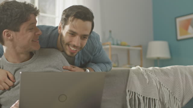 Sweet-Male-Queer-Couple-Spend-Time-at-Home.-Young-Man-Uses-a-Laptop,-His-Partner-Comes-From-Behind-and-Gently-Embraces-Him.-They-Laugh-and-Touch-Hands.-Room-Has-Modern-Interior.