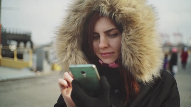 Close-up-shot-of-beautiful-woman-using-smart-phone-technology-app-walking-on-the-street-in-a-warm-jacket-with-a-fluffy-hood