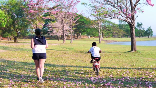 Una-mujer-asiática-gorda-corriendo-en-la-luz-del-sol-natural-por-la-noche,-junto-con-su-hijo-montando-en-bicicleta.--ejercicio-para-una-buena-salud.-Cámara-lenta