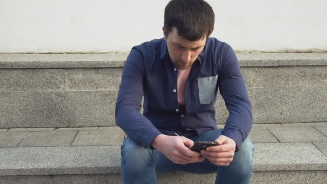 theme-is-man-and-social-networks.-young-Caucasian-sad-male-brunette-in-shirt-is-sitting-in-park-in-Letnec-waiting,-being-late-is-using-hand-to-hold-mobile-phone.-Emotion-resentment-and-disorder
