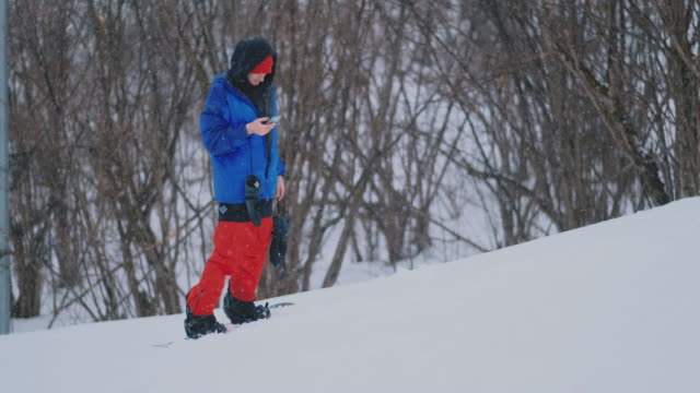 Männlicher-Snowboarder-fährt-das-Board-am-Ski-zur-Schneepiste-und-schreibt-Nachrichten-auf-Smartphone-an-Ihre-Freunde