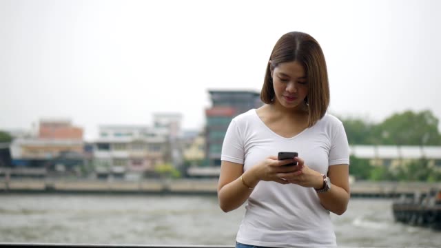 Joven-asiática-usando-el-teléfono-inteligente-disfrutando-de-la-lectura-de-medios-sociales-junto-a-la-valla-en-el-río.