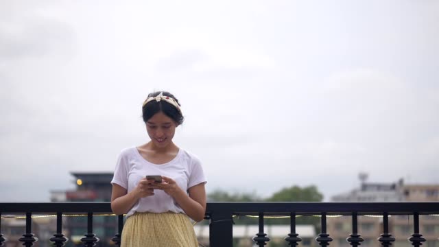 Schöne-junge-asiatische-Frau-mit-Smartphone-genießen-das-Lesen-von-sozialen-Medien-neben-Zaun-am-Fluss.