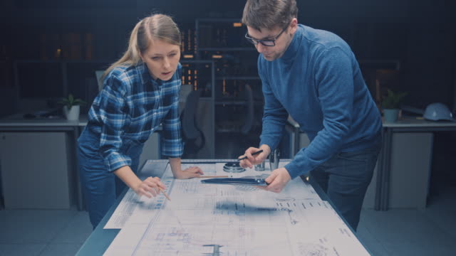 In-der-Dark-Industrial-Design-Engineering-Facility-Male-und-weiblichen-Ingenieurinnen-sprechen-und-arbeiten-Sie-an-einem-Blueprints-Using-Conference-Table.-Auf-dem-Desktop-Zeichnungen,-Entwürfe-und-Elektromotoren-Komponenten,-Teile