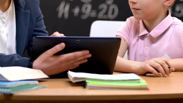 Female-teacher-holding-tablet-PC,-explaining-information-to-little-school-boy