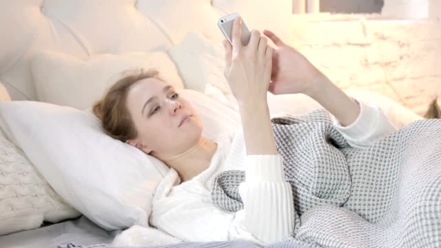 Mujer-joven-que-usa-smartphone-mientras-se-acuesta-en-la-cama
