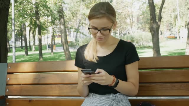Woman-in-glasses-Chatting-On-Mobile-Phone.Relaxed-Girl-Sit-And-Looking-At-Mobile-Phone-In-City-Park.-Beautiful-Woman-Having-Chat-Using-Smartphone-Outdoors.Woman-Chatting-With-Friend.