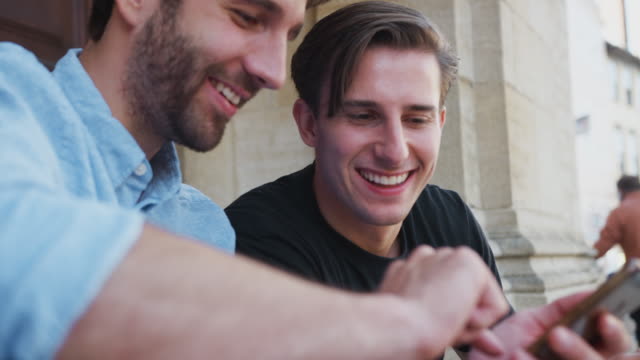 Männliche-Homosexuell-Paar-sitzen-im-Freien-auf-Stufen-des-Gebäudes-Blick-auf-Mobiltelefone