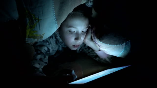 A-child,-a-little-girl,-reads-a-fairy-tale-on-a-tablet-at-night-under-a-blanket-on-the-bed.-Concept-video.-Close-up.-Raw-video.-4K.