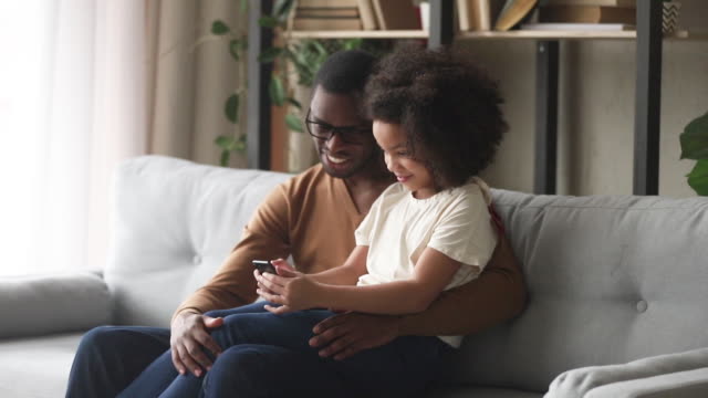 Happy-kid-girl-holding-phone-sit-on-african-dad-lap