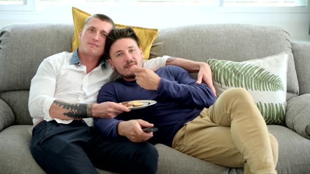 Gay-couple-relaxing-on-couch-watching-tv.-Kissing-on-cheek.