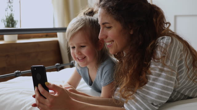Happy-mom-with-kid-daughter-enjoy-using-phone-on-bed