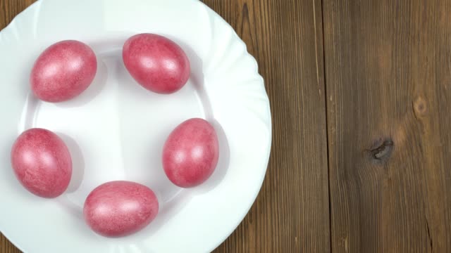 Male-hand-puts-on-the-plate-a-green-Easter-egg-on-the-background-of-red-mother-of-pearl-eggs-in-a-plate,-wooden-background,-copy-space