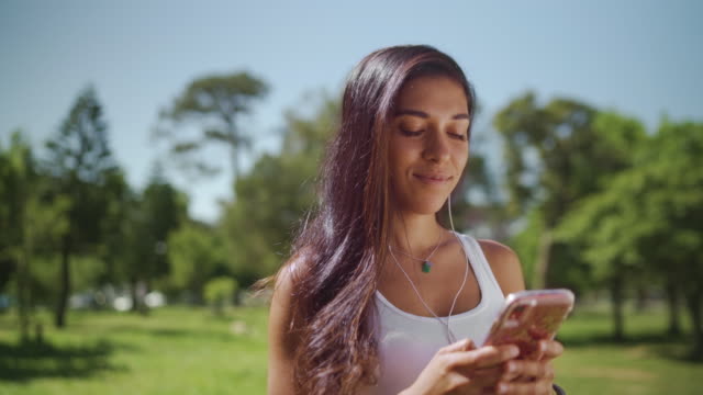 Porträt-einer-glücklichen-brünetten-jungen-Frau,-die-im-Park-mit-Kopfhörern-in-ihren-Ohren-SMS-Nachricht-auf-dem-Smartphone---junge-College-Studentin-auf-ihrem-Handy