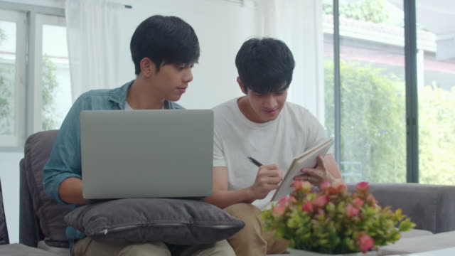 Junge-asiatische-Homosexuell-Paar-arbeiten-Laptop-bei-modernen-haus.-Asia-LGBTQ+-Männer-glücklich-entspannen-Spaß-mit-dem-Computer-und-analysieren-ihre-Finanzen-im-Internet-zusammen,-während-liegen-Sofa-im-Wohnzimmer-im-Haus-Konzept.