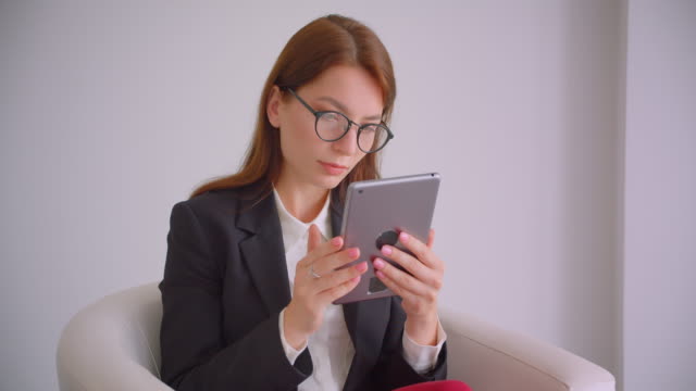 Nahaufnahme-Porträt-der-jungen-kaukasischen-Geschäftsfrau-in-Brille-SMS-auf-dem-Tablet-sitzen-deiner-Sessel-in-der-weißen-Wohnung