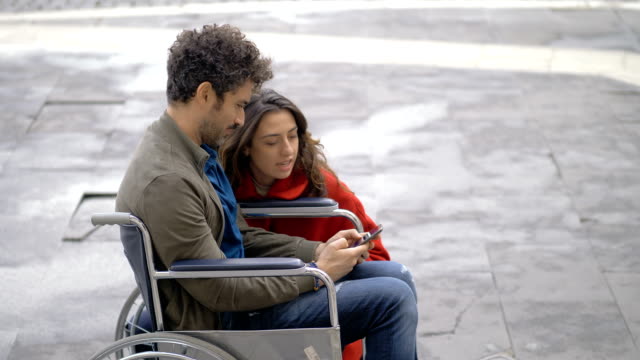 Lächelnder-Mann-im-Rollstuhl-mit-Smartphone-mit-seinem-Freund-auf-der-Straße