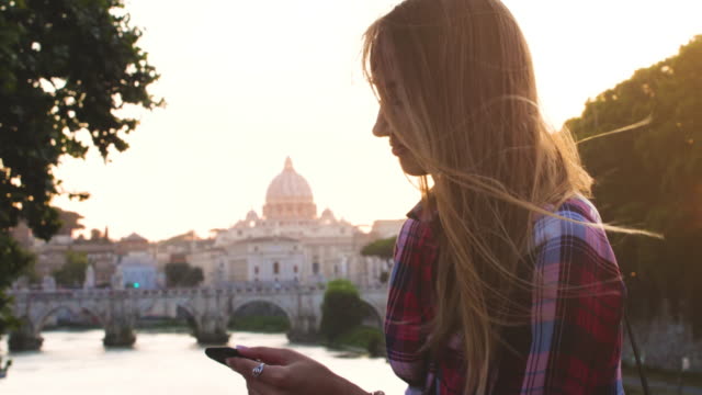 Joven-rubia-turista-sentada-en-un-puente-sobre-el-río-tíber-en-Roma,-utilizando-su-teléfono-inteligente.-Vista-de-San-Pedro-y-la-Ciudad-del-Vaticano,-Italia.
