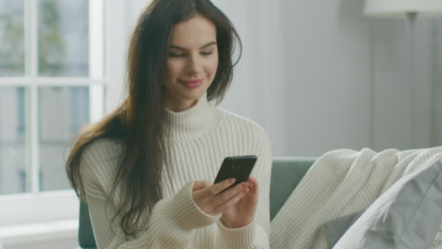 Young-Woman-Using-Smartphone,-Drinks-Tea-while-Sitting-on-the-Chair.-Sensual-Girl-Wearing-Sweater-Surfs-Internet,-Posts-On-Social-Media,-Shares-Pictures-while-Relaxing-in-Cozy-Apartment