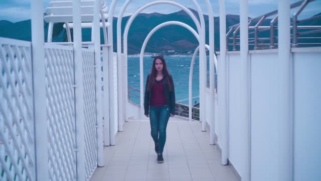 A-beautiful-and-passionate-girl-in-a-biker-jacket-and-jeans-walks-along-the-corridor-of-white-arched-beach-facilities.