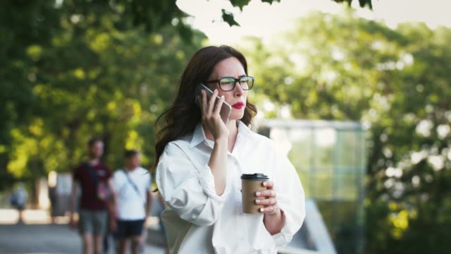 Mädchen-in-Brille-und-weißem-Hemd-sprechen-auf-dem-Smartphone,-halten-Tasse-Kaffee,-eine-Pause-in-Park-mit-grünen-Bäumen.-Geschäftskonzept.-Zeitlupe
