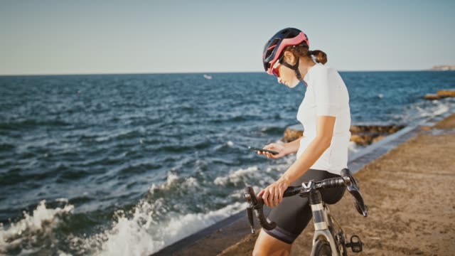 Weibchen-in-Helm-und-Sonnenbrille-sitzt-auf-Trekking-Bike-in-der-Nähe-des-Meeres.-Smartphone-aus-der-Gesäßtasche-ihres-T-Shirts-nehmen-und-Nachricht-eingeben